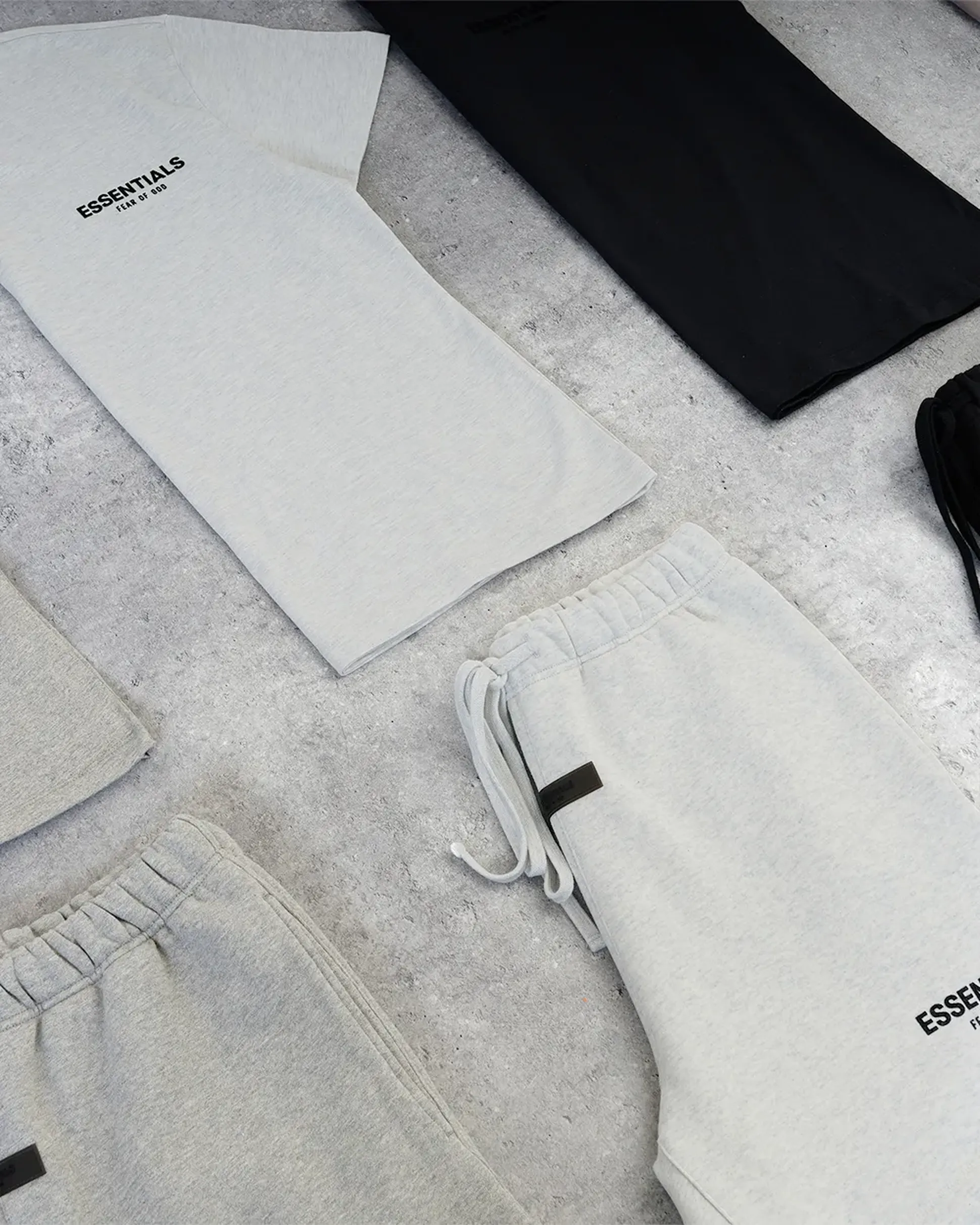 Gray and black "Essentials" branded T-shirts and sweatpants are neatly arranged on a light gray surface.