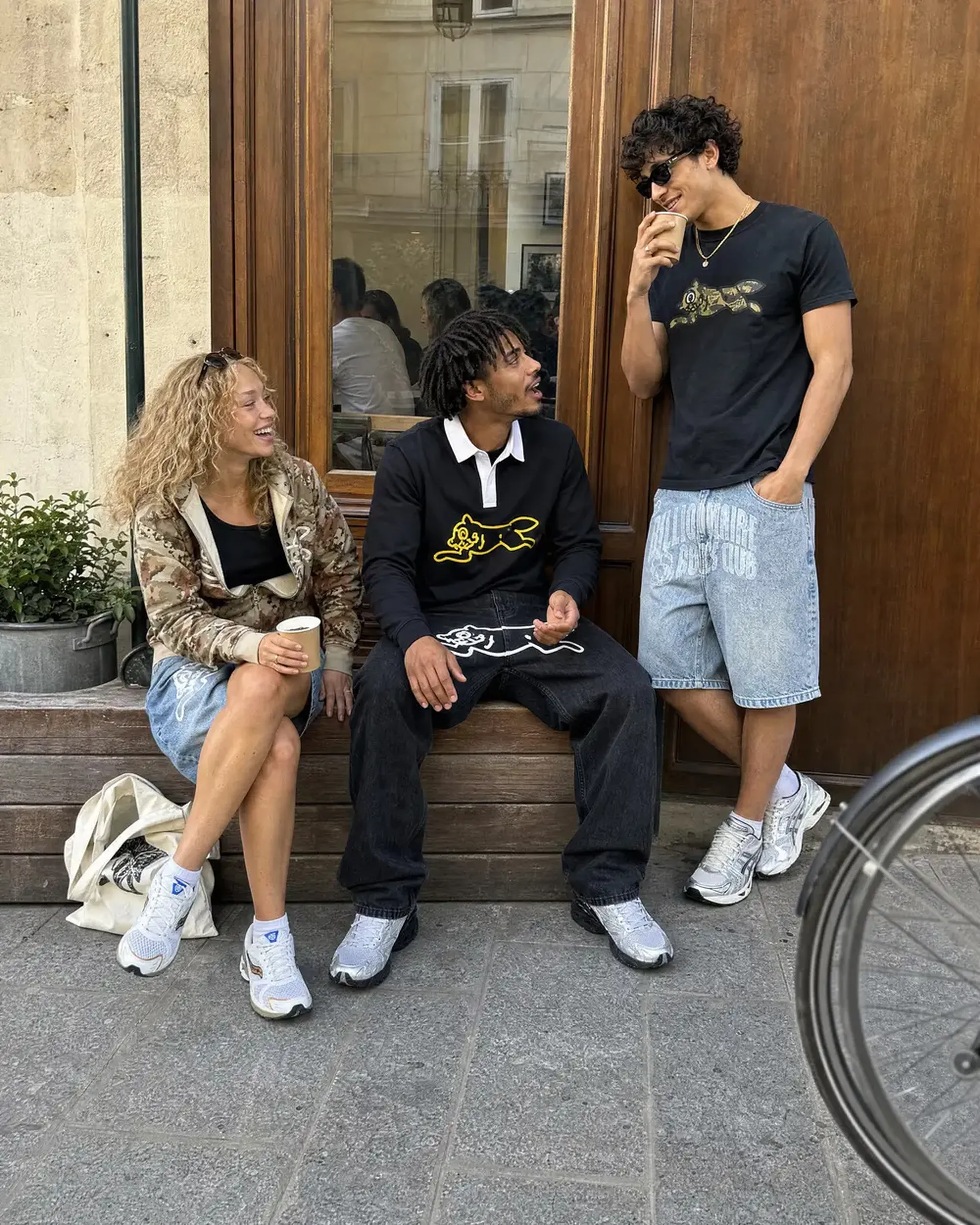 Three people in casual outfits chat and laugh outside a café, with a bicycle wheel visible in the foreground.
