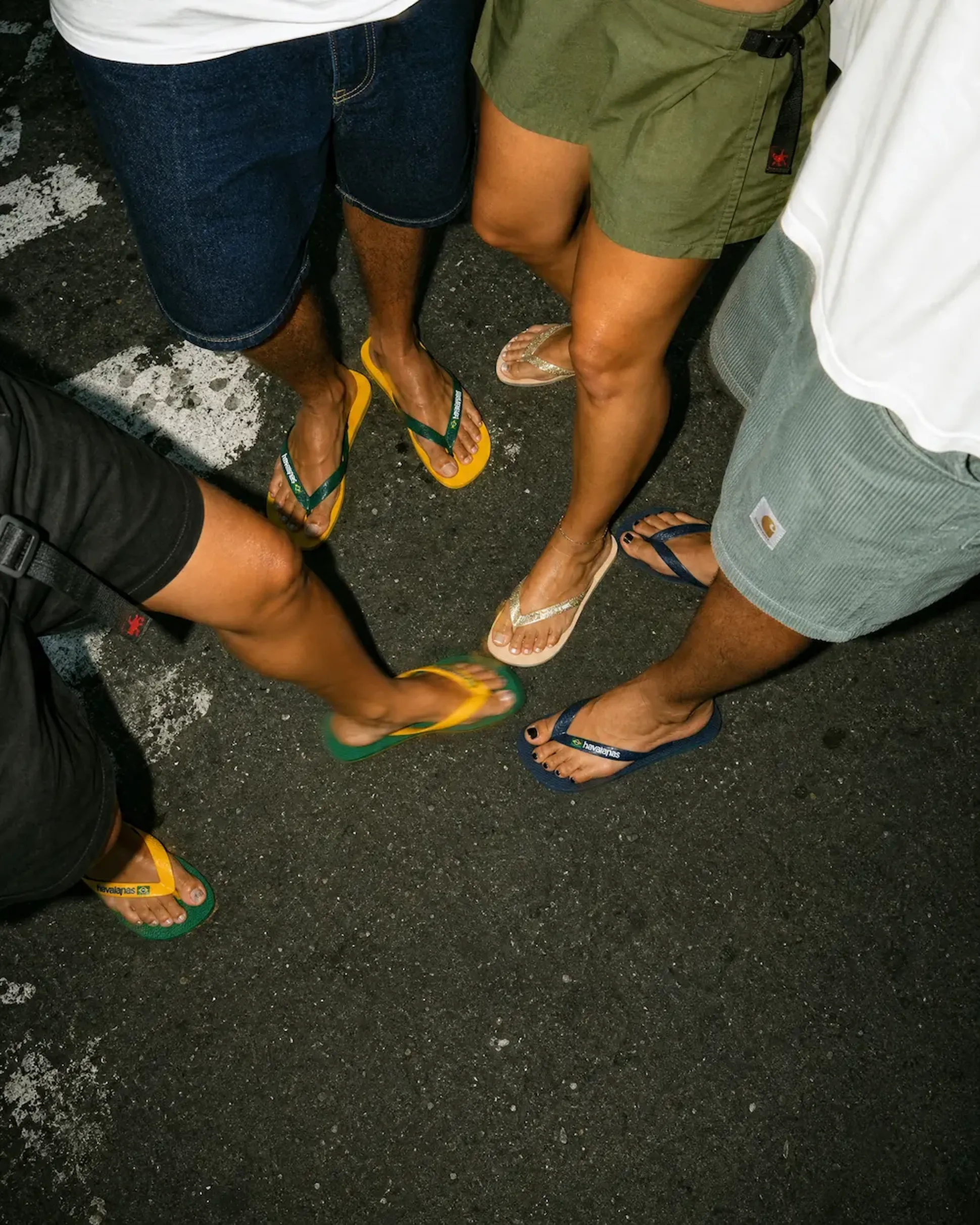 Four people wearing various flip-flops stand on a dark pavement, showcasing casual summer footwear.