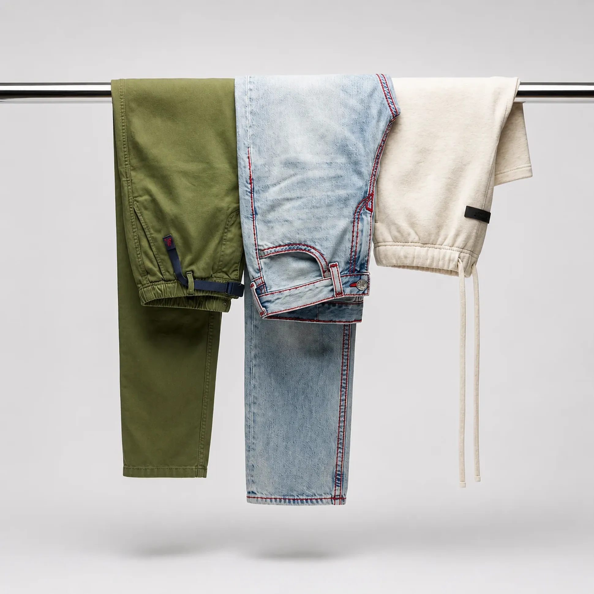 Three pairs of pants hanging on a rod: green cargo, light blue jeans with red stitching, and beige joggers with drawstring.