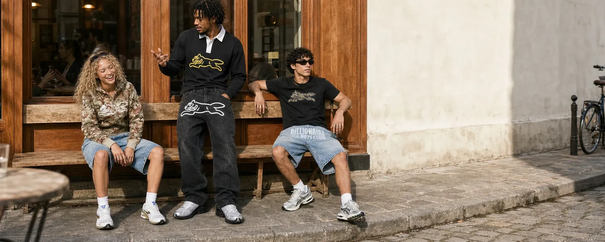 Three people in streetwear sit on a bench outside a cafe, chatting and smiling, with a bicycle parked nearby on the cobblestone street.