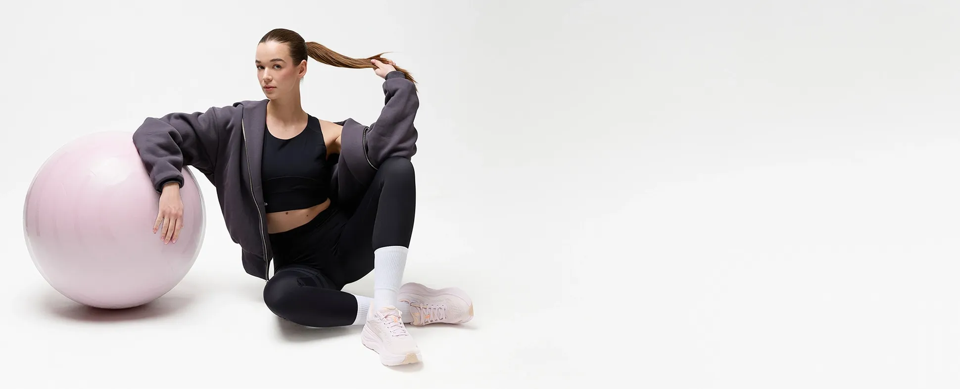 A woman in activewear stretches on a pink exercise ball, raising one leg high. She wears a gray hoodie, black leggings, and pink sneakers.