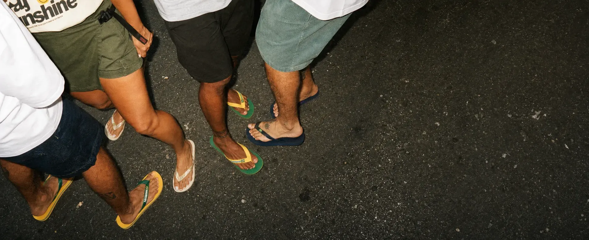 A group of people standing on pavement, wearing colorful flip-flops, shorts, and casual shirts, with visible tattoos and tanned skin.