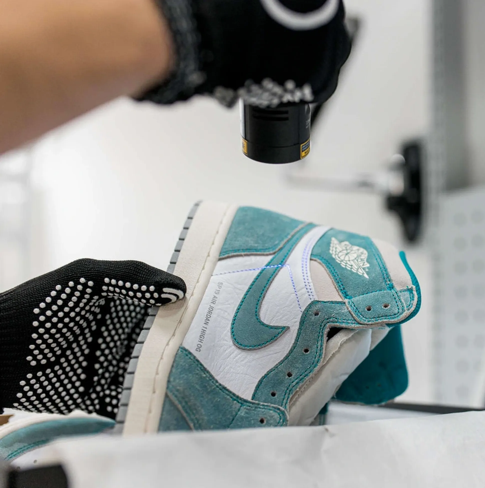 Gloved hand holding a teal and white sneaker with a flashlight, inspecting details in a well-lit room.