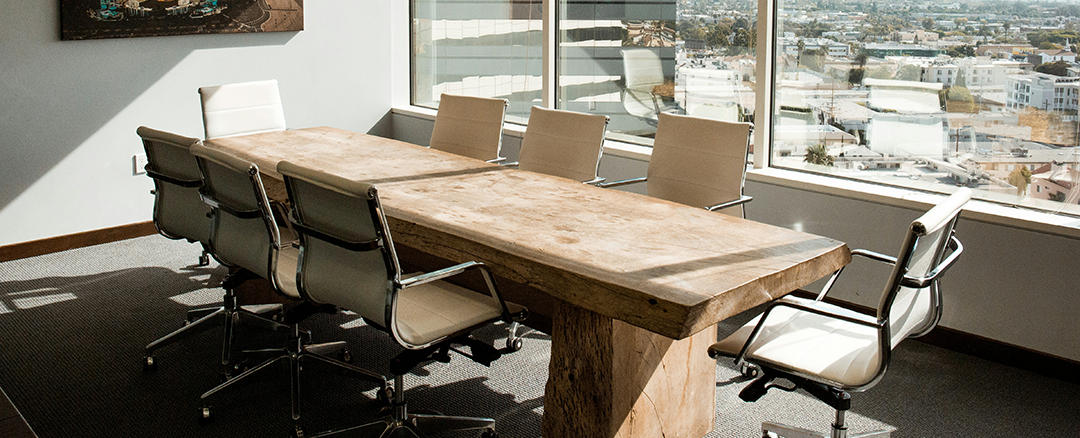 Conference room table A boardroom with a large wooden meeting table surrounded by white office chairs, set in a high-rise space with wide windows overlooking the city.