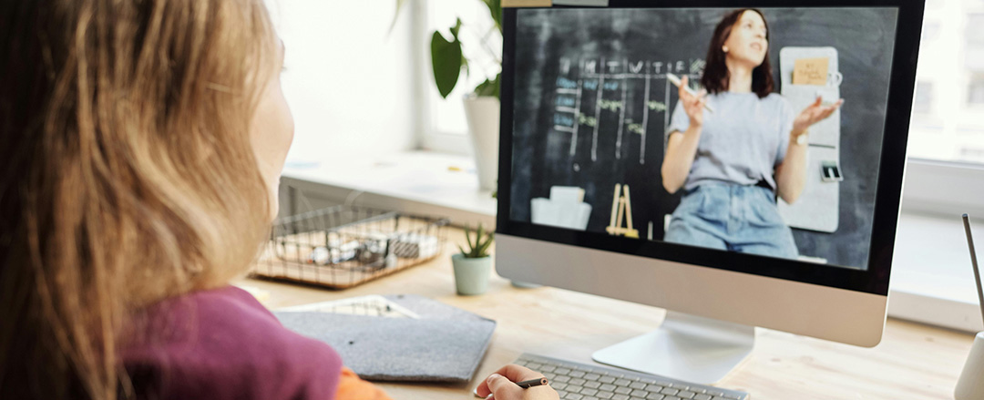 Student watching an online lesson on a computer, taking notes at a desk, illustrating remote learning, hybrid education, and digital classroom access