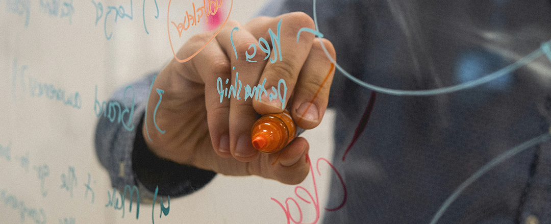 Hand writing glassboard A close-up of a hand using an orange marker to write notes on a transparent glass board, with colorful handwriting and brainstorming elements visible.