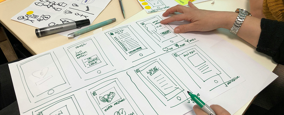 Team collaborating around handwritten wireframes and sketches during a UX design workshop, illustrating human-centered design, collaborative innovation and natural brainstorming.