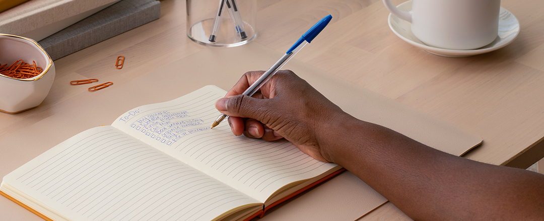 A professional writing a handwritten to-do list in a notebook at a desk with coffee and office supplies, illustrating how handwriting supports focus, productivity, and memory in hybrid work environments.
