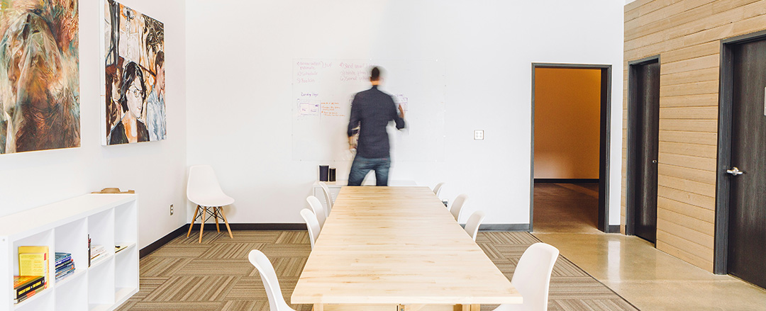 Minimalist collaborative workspace with long wooden meeting table and wall whiteboard used for brainstorming and hybrid team meetings.