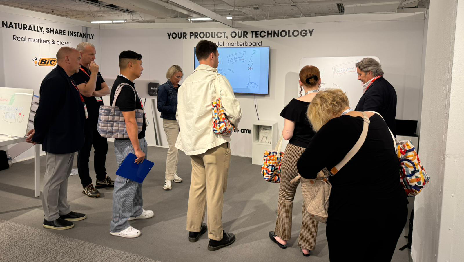 A group of people observes a presentation on a digital whiteboard at a technology booth, with branded bags visible.