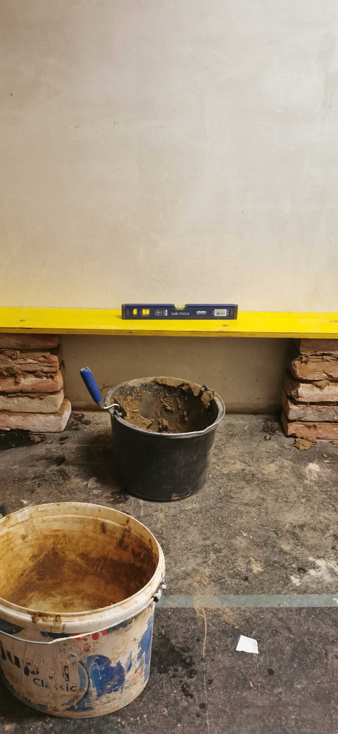Construction setup with yellow wooden slab on bricks, a spirit level on top, and buckets with tools and plaster on a concrete floor. A bench.