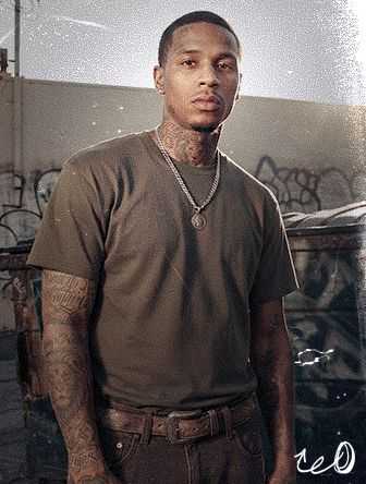 A man with tattoos wearing a dark shirt and chain necklace stands in front of a graffiti-covered wall, looking directly at the camera.