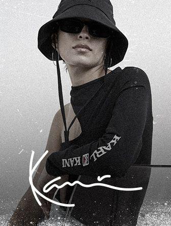 Person wearing a black bucket hat, sunglasses, and a long-sleeve top with "KARL KANI" text, in a grayscale, textured photo.