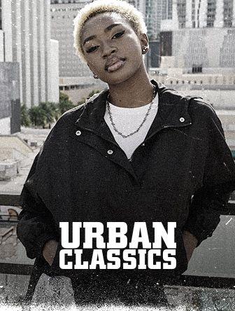 Person in a black jacket stands confidently on a balcony with a cityscape backdrop. Text reads "Urban Classics."