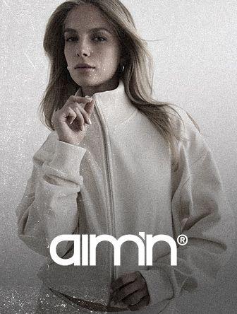 A person wearing a white, high-neck jacket with a calm expression, standing against a neutral background. The text "aimn" is at the bottom.