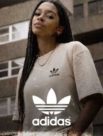 Person wearing a beige Adidas T-shirt with a trefoil logo, standing confidently in an urban environment.