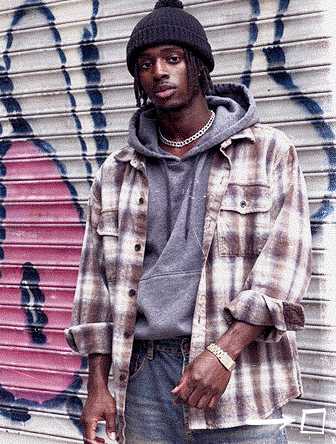 Man in plaid shirt and beanie stands confidently against graffiti-covered metal shutter, wearing a gray hoodie and jeans.
