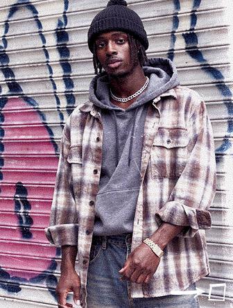 Man in plaid shirt and beanie stands confidently against graffiti-covered metal shutter, wearing a gray hoodie and jeans.