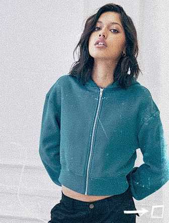 Woman with dark hair wearing a teal zip-up jacket and black pants poses confidently against a light background.