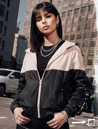 Woman in a two-tone jacket stands confidently on a city street, with buildings in the background. She wears layered necklaces and holds her hands in pockets.