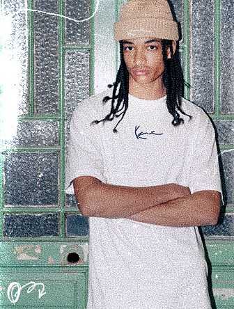 Person in a beanie and white t-shirt with "K" logo stands in front of a textured glass door with arms crossed.