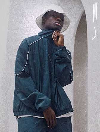 Man in a dark tracksuit and white bucket hat stands pensively, adjusting his collar in front of an archway.