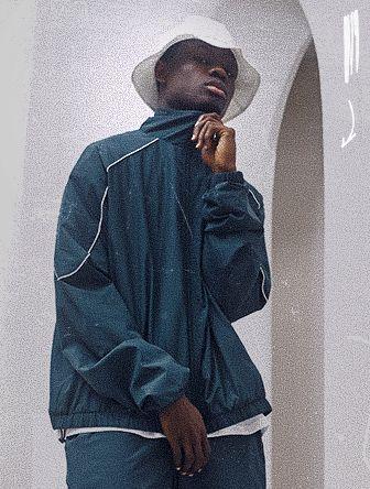 Man in a dark tracksuit and white bucket hat stands pensively, adjusting his collar in front of an archway.