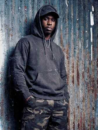 Person wearing a black hoodie and camo pants stands against a corrugated metal wall, looking at the camera with hands in pockets.
