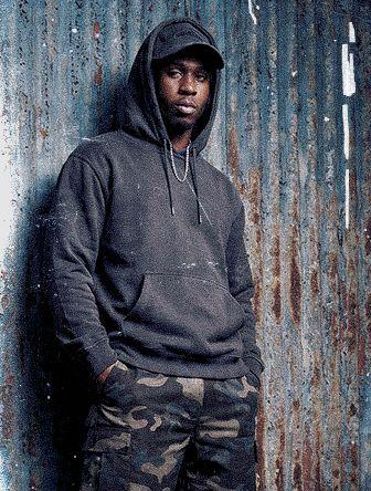 Person wearing a black hoodie and camo pants stands against a corrugated metal wall, looking at the camera with hands in pockets.
