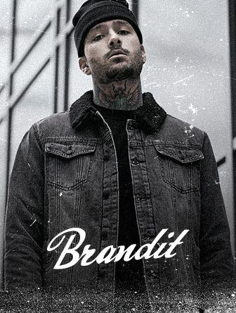 Man in a denim jacket and beanie, with tattoos, stands in front of a building with "Brandit" text overlay.