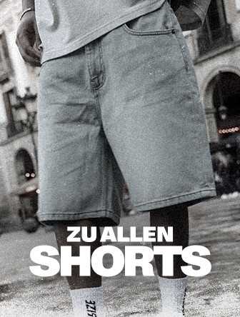 Person wearing knee-length denim shorts and a shirt, standing in a plaza with blurred buildings in the background. Text reads "Zu Allen Shorts."