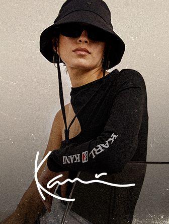 Woman wearing a black bucket hat, sunglasses, and a one-shoulder top with "KARL KANI" text, sitting on a chair.
