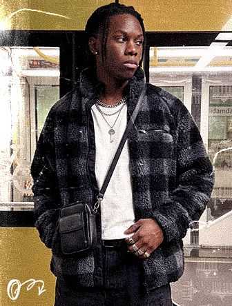 Person in a plaid jacket with a crossbody bag stands in front of a window, wearing layered necklaces and rings, with a yellow background.