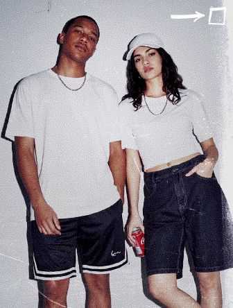 Two people in casual attire pose against a white wall. One holds a soda can, both wearing white tops, with a cap and dark shorts.