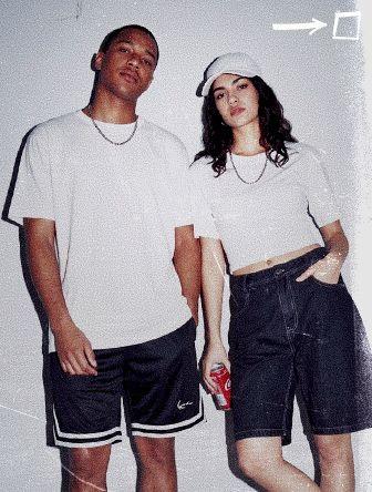Two people in casual attire pose against a white wall. One holds a soda can, both wearing white tops, with a cap and dark shorts.