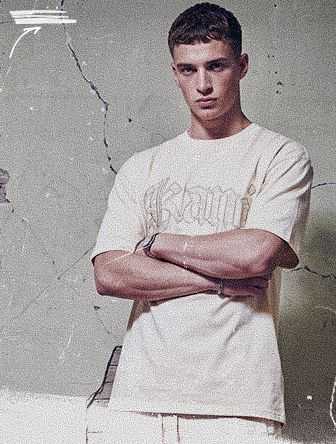 A man in a light-colored T-shirt stands with crossed arms against a textured, cracked wall, looking directly at the camera.