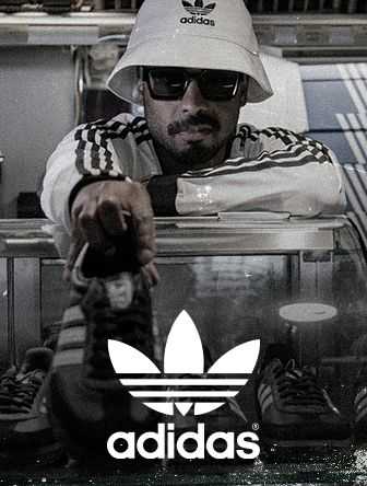 A person wearing a white Adidas hat and sunglasses leans over a glass case filled with Adidas shoes, with the Adidas logo prominently displayed.