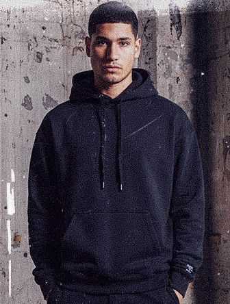 Man wearing a black hoodie stands against a textured, worn concrete wall, hands in pockets, with a serious expression.
