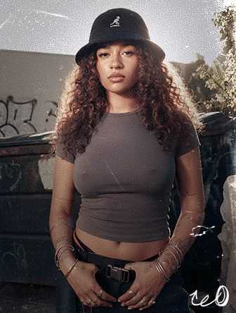 Young woman with curly hair and a bucket hat, wearing a gray crop top, standing confidently in an urban setting with graffiti in the background.