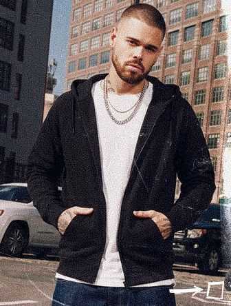 Man standing outdoors in a city, wearing a black hoodie and white shirt, with hands in pockets, against a backdrop of tall buildings and parked cars.