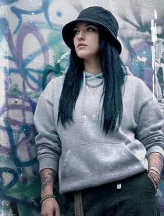 A woman with long dark hair wearing a gray hoodie and black bucket hat stands in front of a graffiti-covered wall, looking to the side.