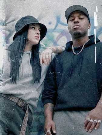 Two individuals in streetwear stand against a graffiti wall; one woman in a bucket hat and hoodie rests her arm on a man's shoulder.