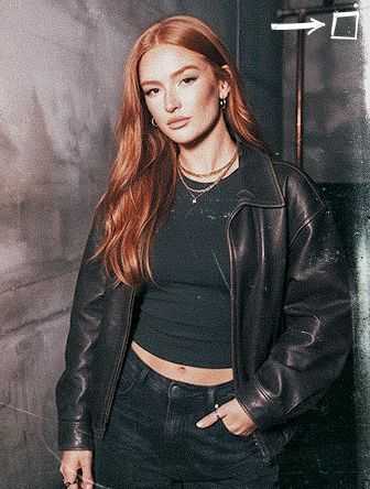 A person with long red hair wearing a black jacket and crop top stands against a textured wall, looking into the camera.