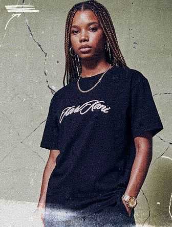 Person with long braids wearing a black T-shirt with script text, standing against a textured wall.