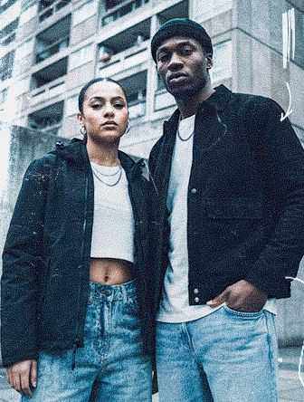 A man and woman stand confidently in casual clothing against an urban concrete building backdrop.