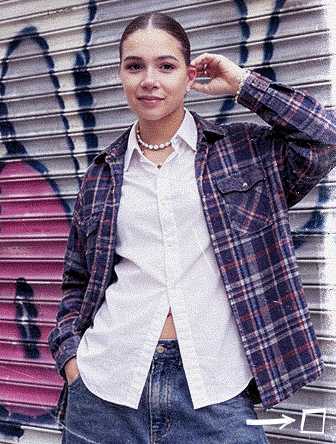 Person in a plaid shirt and jeans stands confidently in front of a graffiti-covered metal shutter.