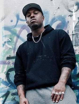 A person in a black hoodie and cap stands confidently in front of a graffiti-covered wall, wearing silver chains and rings.