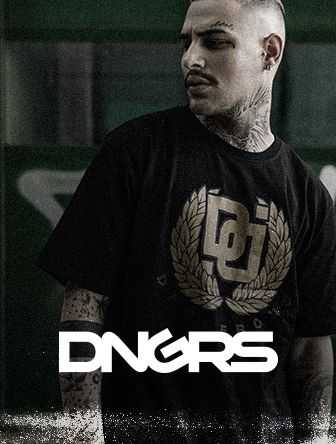 Man with tattoos wearing a black DNGRS shirt and looking sideways, standing against a green background.
