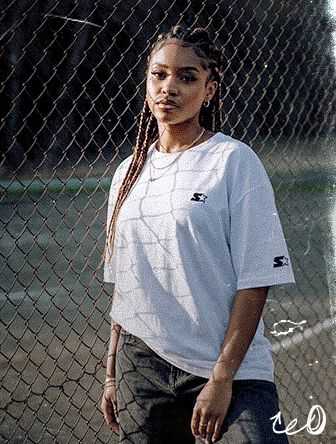 Woman with braided hair wearing a white t-shirt stands in front of a chain-link fence, looking at the camera.